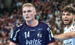 Zwei männliche Handballspieler auf dem Spielfeld. Der eine, in einem blauen Trikot mit der Nummer 14, wirkt konzentriert. Der andere, in einem weißen Trikot, hat den Mund offen und legt eine Hand auf die Schulter des blauen Spielers. Im Hintergrund ist eine verschwommene Zuschauermenge zu sehen.