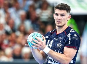 Ein männlicher Handballspieler in einem marineblauen Trikot hält einen blau gemusterten Ball in der Hand und ist während des Spiels voll bei der Sache. Im Hintergrund sind verschwommene Zuschauer zu sehen.