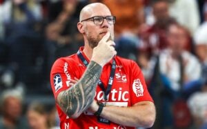 Ein kahlköpfiger Mann mit Brille und Tattoos, der ein rotes Sportshirt trägt, steht da, die Hand am Kinn, und wirkt nachdenklich. Im Hintergrund sind verschwommene Menschen und Stadionplätze zu sehen.