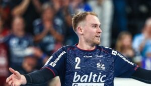 Ein männlicher Handballspieler in einem blauen Trikot mit der Aufschrift „Baltic“ auf der Vorderseite steht während eines Spiels mit ausgestreckten Armen da. Im Hintergrund ist eine verschwommene Menschenmenge zu sehen.