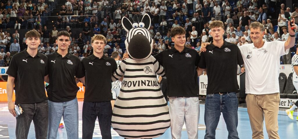 27_Ehrung_EM_Junioren_Titel Sechs Personen – fünf junge Männer in einheitlichen schwarzen Hemden und Jeans sowie ein älterer Mann in einem weißen Hemd – stehen lächelnd neben einer Person in einem Zebra-Maskottchenkostüm auf einem Sportplatz, im Hintergrund jubelnde Zuschauer.