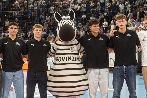 Sechs Personen – fünf junge Männer in einheitlichen schwarzen Hemden und Jeans sowie ein älterer Mann in einem weißen Hemd – stehen lächelnd neben einer Person in einem Zebra-Maskottchenkostüm auf einem Sportplatz, im Hintergrund jubelnde Zuschauer.