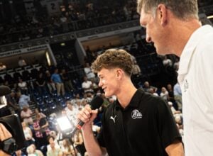Ein junger Mann in einem schwarzen Sportshirt spricht bei einer Hallenveranstaltung in ein Mikrofon, neben ihm steht ein lächelnder älterer Mann. Im Hintergrund beobachtet eine Menschenmenge das Geschehen von den Tribünen aus.