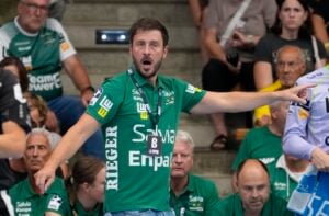 Ein Mann in einem grünen Sport-T-Shirt steht während einer Hallensportveranstaltung da, zeigt mit dem Finger auf etwas und ruft laut. Mehrere sitzende Personen in grünen T-Shirts und Zuschauer im Hintergrund verfolgen das Geschehen.