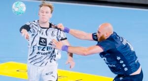 Zwei männliche Handballspieler kämpfen um den Ball; der Spieler in Weiß hält den Ball in der Hand, während der Spieler in Blau nach ihm greift und ihn am Trikot packt. Beide wirken während dieser intensiven Spielszene auf einem blauen Hallenfeld hochkonzentriert.