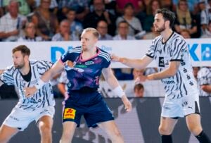 Drei männliche Handballspieler sind auf dem Spielfeld im Einsatz, zwei davon tragen weiß gestreifte Trikots und einer ein blau-rosa Trikot. Im Hintergrund ist die Zuschauermenge verschwommen zu sehen, die das Spiel gebannt verfolgt.
