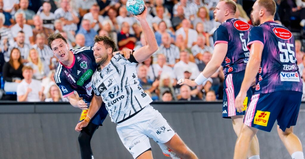 HANDBALL 2026/27: THW Kiel - TSV Hannover-Burgdorf Ein Handballspieler in weißer Spielkleidung versucht während eines Spiels einen Wurf, während er von Spielern in blauen Trikots verteidigt wird; im Hintergrund ist eine Zuschauermenge zu sehen.