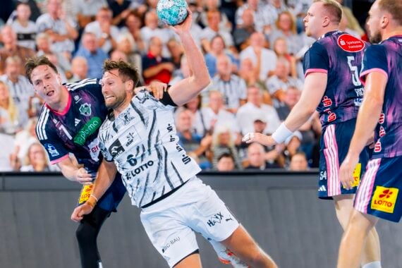 Ein Handballspieler in weißer Spielkleidung versucht während eines Spiels einen Wurf, während er von Spielern in blauen Trikots verteidigt wird; im Hintergrund ist eine Zuschauermenge zu sehen.