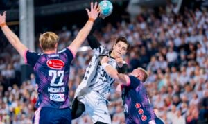 Ein Handballspieler im weißen Trikot springt zum Wurf hoch, während zwei Gegner in blauen Trikots die Arme heben, um ihn während eines spannenden Spiels in einer vollbesetzten Halle zu blocken.