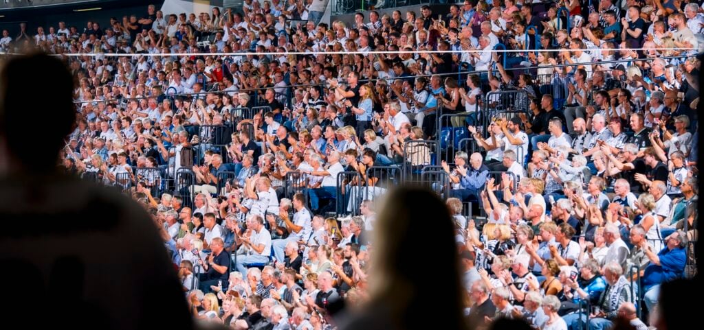 HANDBALL 2026/27: THW Kiel - TBV Lemgo Lippe Eine große Menschenmenge sitzt auf den Stadionplätzen, klatscht und verfolgt eine Veranstaltung. Im Vordergrund sind zwei Personen zu sehen, verschwommen und als Silhouetten.