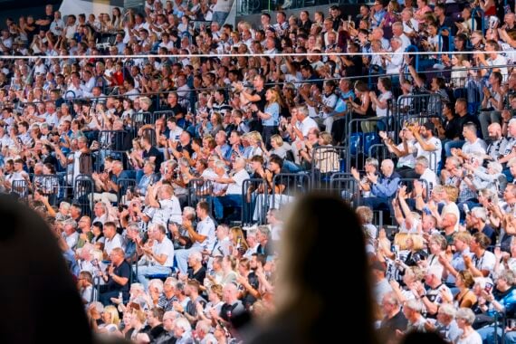 Eine große Menschenmenge sitzt auf den Stadionplätzen, klatscht und verfolgt eine Veranstaltung. Im Vordergrund sind zwei Personen zu sehen, verschwommen und als Silhouetten.