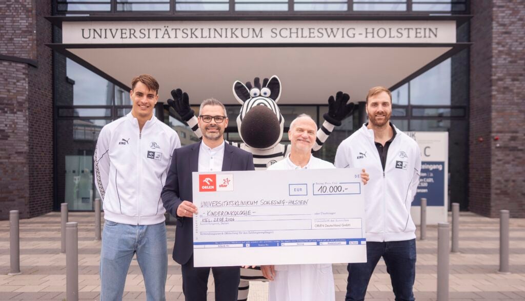 27_UKSH_ORLEN_Koester Vier Personen und ein Zebra-Maskottchen stehen vor dem Universitätsklinikum Schleswig-Holstein, halten einen großen symbolischen Scheck über 10.000 Euro in den Händen und lächeln in die Kamera.