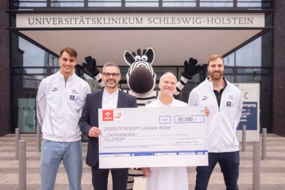 Vier Personen und ein Zebra-Maskottchen stehen vor dem Universitätsklinikum Schleswig-Holstein, halten einen großen symbolischen Scheck über 10.000 Euro in den Händen und lächeln in die Kamera.