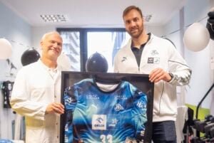 Zwei lächelnde Männer halten ein gerahmtes blaues Sporttrikot mit der Nummer 33 in den Händen. Im Hintergrund sind schwarz-weiße Luftballons und Dekorationen zu sehen, was auf eine Feier hindeutet.