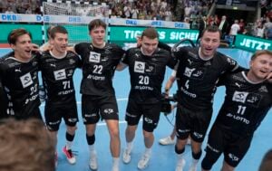 Sechs männliche Handballspieler in schwarzen Trikots stehen Arm in Arm und feiern auf einem blauen Hallenfeld, während im Hintergrund eine jubelnde Menge zu sehen ist.