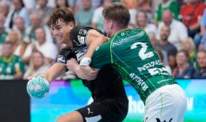 Zwei männliche Handballspieler kämpfen während eines Spiels um den Ball; dabei versucht einer in schwarzem Trikot, sich von einem Spieler in grünem Trikot zu lösen, während im Hintergrund eine Zuschauermenge zusieht.