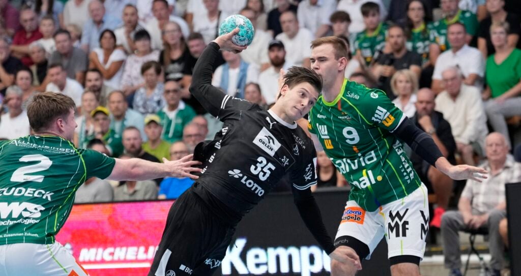 2609_FAG-Kiel_11056 Ein Handballspieler in einem schwarzen Trikot bereitet sich darauf vor, den Ball zu werfen, während er von zwei Spielern in grünen Trikots verteidigt wird. Im Hintergrund verfolgen die Zuschauer das Geschehen gespannt von den Tribünen aus.