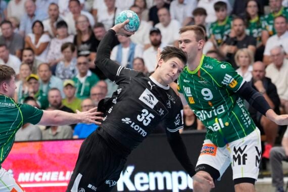 Ein Handballspieler in einem schwarzen Trikot bereitet sich darauf vor, den Ball zu werfen, während er von zwei Spielern in grünen Trikots verteidigt wird. Im Hintergrund verfolgen die Zuschauer das Geschehen gespannt von den Tribünen aus.