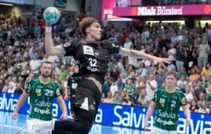 Ein Handballspieler in schwarzer Spielkleidung springt hoch, um einen Wurf auszuführen, während zwei gegnerische Spieler in grüner Spielkleidung zusehen. Die Halle ist voll mit Zuschauern, und im Hintergrund sind Werbebanner zu sehen.