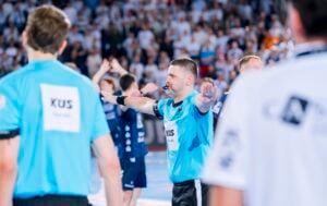 Ein Schiedsrichter in einem hellblauen Trikot gibt während eines Hallenhandballspiels mit ausgestreckten Armen ein Zeichen, im Hintergrund sind Spieler und eine verschwommene Zuschauermenge zu sehen.