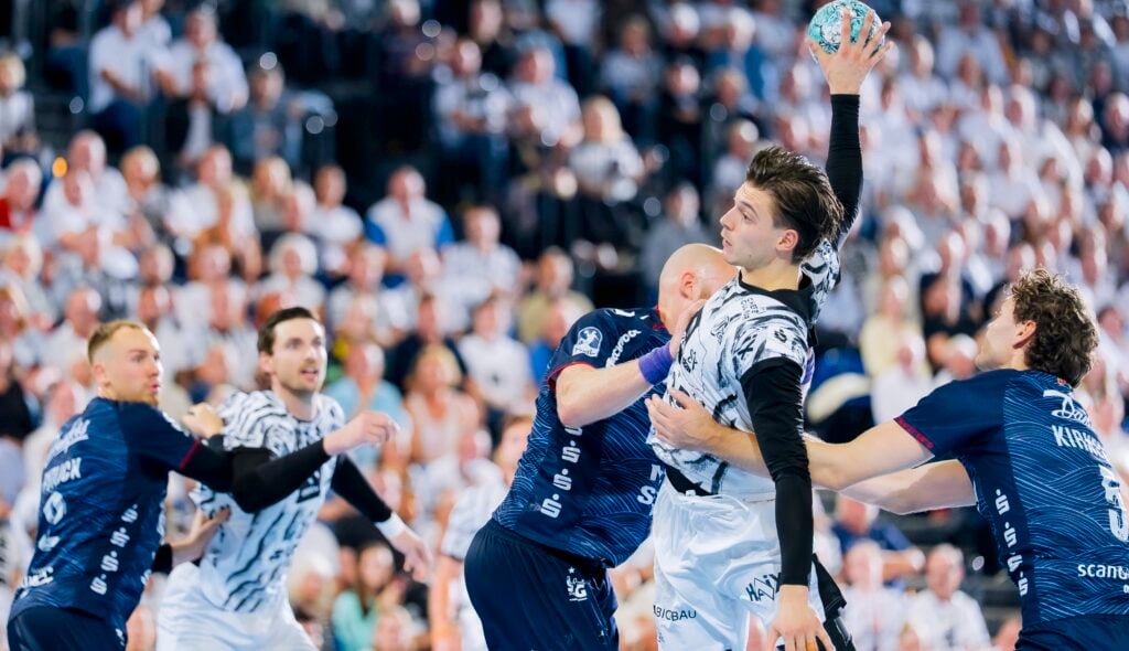 Ein Handballspieler in Weiß springt hoch, um den Ball zu werfen, während er von Spielern in blauen Trikots während eines spannenden Spiels verteidigt wird; im Hintergrund ist eine große Zuschauermenge zu sehen.