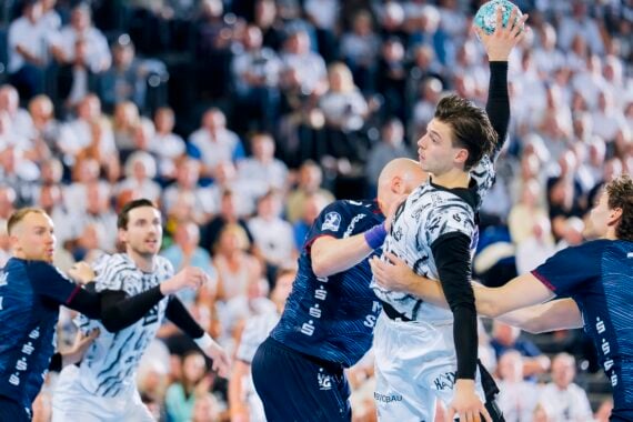 Ein Handballspieler in Weiß springt hoch, um den Ball zu werfen, während er von Spielern in blauen Trikots während eines spannenden Spiels verteidigt wird; im Hintergrund ist eine große Zuschauermenge zu sehen.