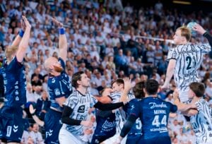 Ein Handballspieler in einem weiß gestreiften Trikot springt hoch, um einen Wurf zu versuchen, während mehrere Verteidiger in dunkelblauen Trikots die Arme heben, um ihn zu blocken. Auf dem Spielfeld herrscht Gedränge, im Hintergrund ist eine große Zuschauermenge zu sehen.