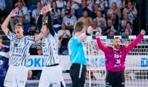 Zwei Handballspieler in weißen Trikots feiern mit erhobenen Armen, während ein Schiedsrichter in Blau ein Zeichen gibt. Auch ein Torwart in einem rosa Trikot jubelt in der Nähe des Tors. Im Hintergrund schaut das Publikum zu – eine Szene, wie sie auch am Samstag beim Handball-Derby zwischen dem THW Kiel und der SG Flensburg-Handewitt zu sehen sein könnte.