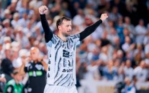 Ein Handballspieler in einem schwarz-weißen Trikot reckt jubelnd beide Arme in die Höhe; im Hintergrund sind eine verschwommene Menschenmenge und der Trainer zu sehen.