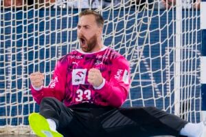 Ein Handballtorwart in einem rosa Trikot mit der Nummer 33 sitzt auf dem Spielfeld vor dem Tor und jubelt leidenschaftlich mit geballten Fäusten und weit geöffnetem Mund.
