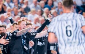 Ein Trainer mit roten Haaren ruft leidenschaftlich und gestikuliert mit geballten Fäusten während einer gut besuchten Hallensportveranstaltung – ein Vorgeschmack auf die mitreißende Atmosphäre, die am Samstag erwartet wird, wenn das Handball-Derby zwischen dem THW Kiel und der SG Flensburg-Handewitt stattfindet. Im Vordergrund ist ein Spieler in einem zebra-gestreiften Trikot mit der Nummer 6 zu sehen, der sich voll und ganz auf das Geschehen konzentriert.