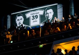 Die Zuschauer sitzen unter einem großen Schild, auf dem zwei gezeichnete Gesichter und ein Trikot mit der Aufschrift „THW Kiel 39 Jicha“ zu sehen sind, das von einer dramatischen Beleuchtung in der Halle in Szene gesetzt wird, in der der THW Kiel das Heimspiel gegen Hannover-Burgdorf mit 29:26 gewinnt.