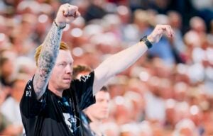 Ein Trainer mit Tattoos hebt während einer Sportveranstaltung beide Arme, um ein Signal zu geben. Er trägt ein schwarzes Hemd und eine Armbanduhr, und im Hintergrund ist eine verschwommene Menschenmenge zu sehen, während der THW Kiel in der Handball-Bundesliga beim ThSV Eisenach antritt.