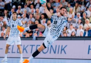 Ein männlicher Handballspieler in einem schwarz-weißen Trikot springt in die Luft, um den Ball zu werfen. Im Hintergrund sind ein weiterer Spieler und Zuschauer zu sehen. Die Szene ist spannend und voller Action.
