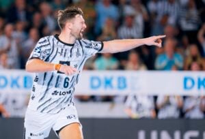 Ein männlicher Handballspieler in einem schwarz-weißen Trikot jubelt während eines Spiels, zeigt mit dem Finger auf etwas und ruft mit einem Lächeln. Im Hintergrund sind eine verschwommene Menschenmenge und ein deutlich erkennbares DKB-Logo zu sehen.
