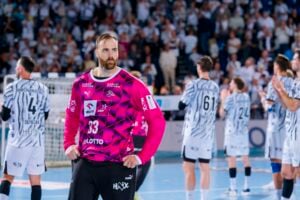 Im Mittelpunkt steht ein bärtiger Handballspieler in einem knallpinken Trikot, der die Hände in die Hüften gestemmt hat, während sich im unscharfen Hintergrund auf einer Halle andere Spieler in schwarz-weißen Trikots versammeln und klatschen.