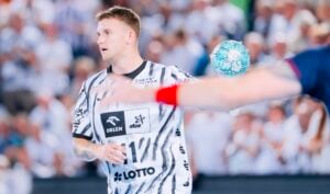 Ein Handballspieler in einem schwarz-weißen Trikot steht auf dem Spielfeld, während ein türkisfarbener Ball in der Luft schwebt. Im Vordergrund ist der Arm eines anderen Spielers zu sehen, der ein rotes Armband trägt. Im Hintergrund ist ein verschwommenes Publikum zu erkennen.
