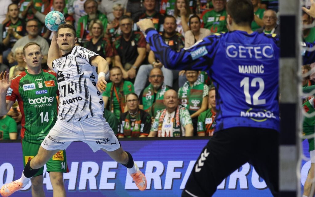 Ein Handballspieler im weißen Trikot springt hoch, um zu werfen, während er dem Torwart in Blau gegenübersteht. Im Hintergrund sind Zuschauer in grünen Trikots zu sehen, die das spannende Geschehen auf dem Spielfeld verfolgen.