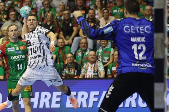 Ein Handballspieler im weißen Trikot springt hoch, um zu werfen, während er dem Torwart in Blau gegenübersteht. Im Hintergrund sind Zuschauer in grünen Trikots zu sehen, die das spannende Geschehen auf dem Spielfeld verfolgen.