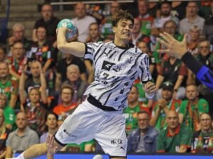 Ein Handballspieler in einem schwarz-weißen Trikot springt in der Luft, um den Ball auf das Tor zu werfen, während im Hintergrund eine Menge Zuschauer gespannt zuschaut.
