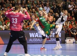 Ein Handballspieler in grün-roter Kleidung versucht, einen Wurf anzusetzen, während zwei Gegner in weißen Trikots verteidigen. Der Torwart in rosa beobachtet das Geschehen. Im Hintergrund ist eine Zuschauermenge zu sehen.