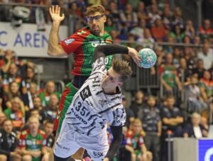 Ein Handballspieler in Weiß wirft den Ball, während ein Verteidiger in Grün und Rot versucht, ihn zu blocken. Das Publikum verfolgt das spannende Geschehen in der Sporthalle.