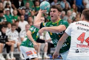 Ein Handballspieler in einem grünen Trikot macht sich bereit, den Ball zu werfen, während Gegner in weißen Trikots verteidigen. Im Hintergrund beobachten die Zuschauer das Geschehen gespannt.