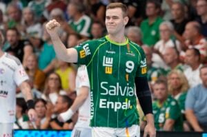 Ein Handballspieler in einem grünen Trikot mit der Nummer 9 reckt die Faust in die Höhe, während er auf dem Spielfeld steht und im Hintergrund eine Menge Zuschauer zu sehen ist.