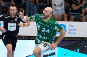 Ein Handballspieler in einem grünen Trikot mit der Nummer 24 feiert mit erhobener Faust. Im Hintergrund ist ein weiterer Spieler in einem schwarzen Trikot zu sehen, und auf den Tribünen sind Zuschauer zu erkennen.
