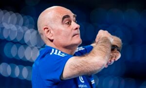 Ein kahlköpfiger Mann in einem blauen Sportshirt gestikuliert mit den Armen und wirkt konzentriert – vielleicht bereitet er sich gerade darauf vor, dass der THW Kiel am Samstag in der Merkur Ostseehalle auf den TSV Hannover-Burgdorf trifft. Der Hintergrund ist durch blaue und weiße kreisförmige Lichter verschwommen.