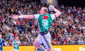 Ein Handballspieler in einem grün-weißen Trikot springt während eines Spiels hoch, um den Ball zu werfen, während im Hintergrund eine große Zuschauermenge zuschaut. Am Samstag trifft der THW Kiel in der Merkur Ostseehalle auf den TSV Hannover-Burgdorf.