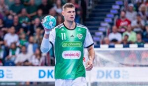 Ein männlicher Handballspieler in einem grün-weißen Trikot hält einen blaugrünen Ball auf einem Hallenfeld in der Hand, während im Hintergrund Zuschauer und ein Tor verschwommen zu sehen sind.