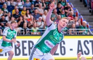 Ein Handballspieler in einem grün-weißen Trikot jubelt und zeigt mit einem Finger nach oben, während im Hintergrund eine jubelnde Menschenmenge zusieht.