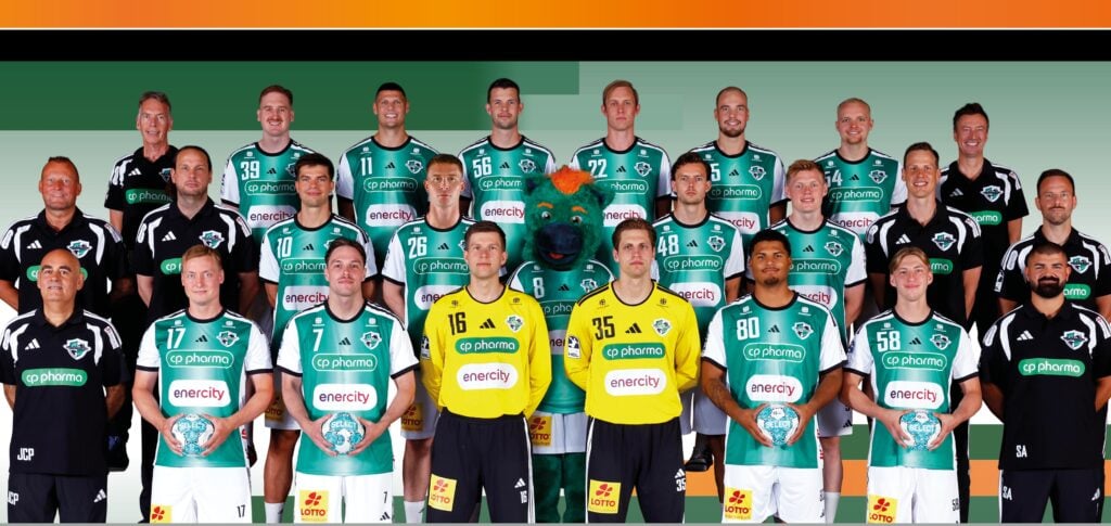 Eine Herren-Handballmannschaft posiert in grünen Trikots, wobei zwei Torhüter in gelben Trikots vorne in der Mitte stehen. Ein Maskottchen in einem Bärenkostüm steht in der Mitte. Trainer und Betreuer stehen an den Seiten.